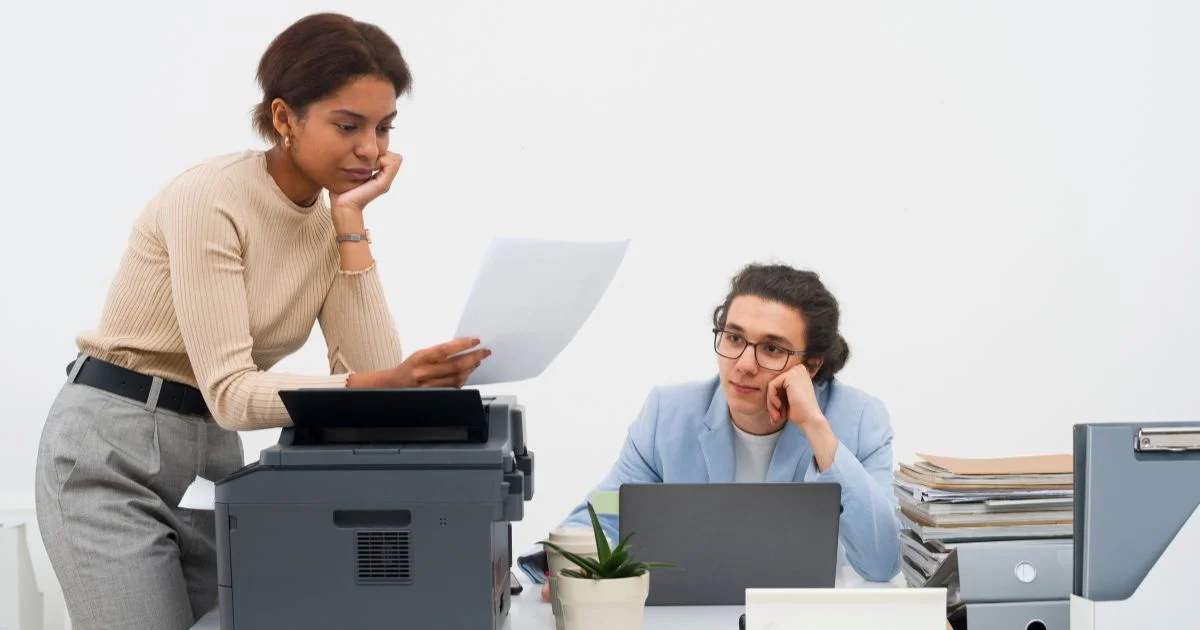 Two coworkers reviewing a printed document beside an office printer.